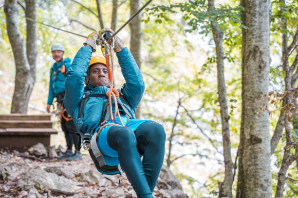 Black male engaging in an outdoor zipline activity. The image features a young adult wearing a helmet and harness, displaying focus and enjoyment. The background shows another participant preparing for the thrilling experience in a natural, wooded setting.