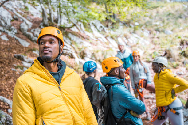 Male and female adventurers of mixed ethnicities gearing up in protective helmets and harnesses for a zipline activity in a forested landscape. The group displays a sense of anticipation and focus dressed in casual outdoor attire, ready for an adventurous day in nature.