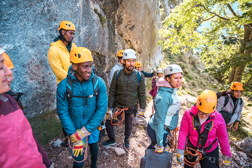 A diverse group of male and female adventurers wearing helmets and harnesses prepares for a zipline adventure in a natural, rocky setting. They appear focused and equipped for an adrenaline-filled experience, with safety gear prominently displayed.