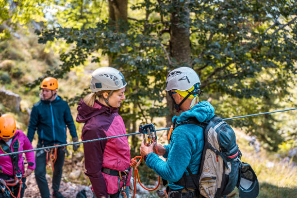 A diverse group of adults are seen preparing for a thrilling zipline experience outdoors. The instructor, wearing professional safety gear, is securing the harness of a female participant. The setting is lush with greenery, suggesting an adventurous outdoor activity. Everyone is equipped with helmets and practical attire, ready for the zipline adventure.