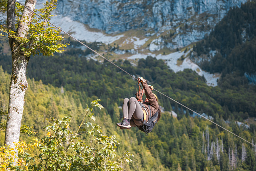 An energetic female enjoys a thrilling zip line journey over scenic mountain forests. Wearing safety gear and casual outdoor attire, she experiences the exhilaration of aerial travel in a natural setting.