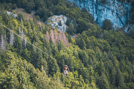 A female engaging in an exhilarating zip line adventure across a dense forest canopy. She is wearing safety gear and appears mid-descent on the cable, with a scenic view of rocky cliffs and greenery in the background.