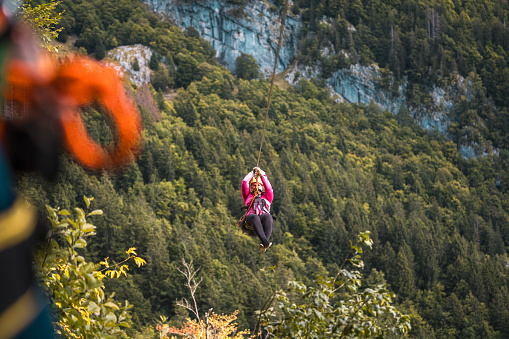 A female in a pink top and safety gear enjoys a thrilling zip line ride above a dense forest canopy. The lush green trees and mountainous backdrop suggest an outdoor adventure activity.