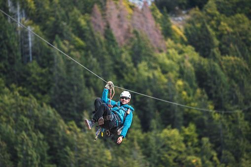 An adult male in a protective helmet and harness enjoys a zipline adventure, soaring above a lush forest. His attire is casual, suitable for outdoor activities. The scene highlights a thrilling aerial pursuit in a natural setting.