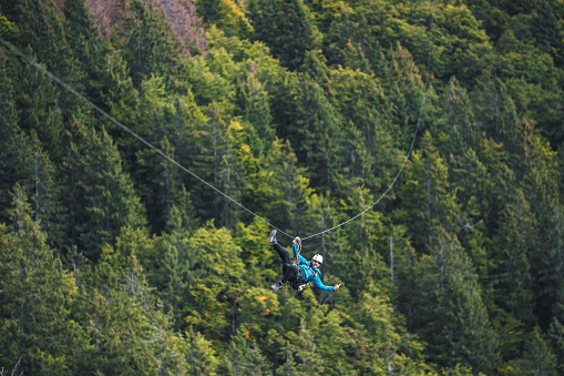 Young adult male engaging in a zip line adventure, soaring above a lush green forest. He is wearing safety gear and a helmet, showing excitement and thrill, embodying the spirit of outdoor adventure sports.