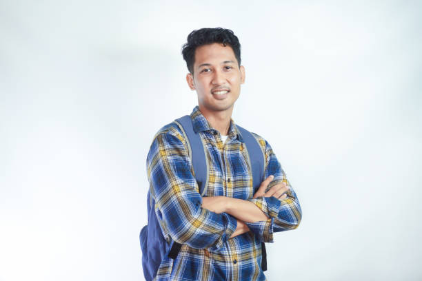 Smiling confident young Asian man student wearing casual clothes and backpack folded hands and looking on camera isolated on white background. high school university college concept