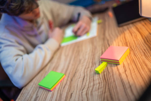 A focused Caucasian teenage boy is captured arranging colorful sticky notes on a wooden table indoors. He wears casual clothes and uses adhesive tape for his study session, creating an organized workspace at home.