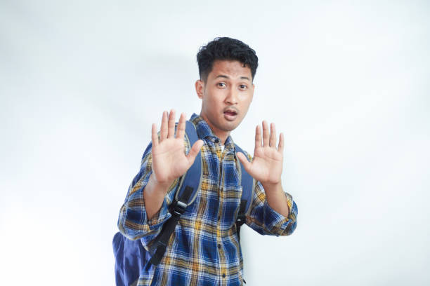 istock Scared young Asian student man in casual clothes with backpack makes stop gesture and tries to protect herself from something isolated on white background. High school university college concept 2054815496