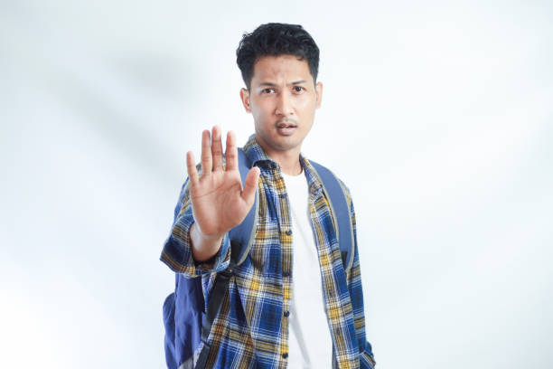 istock Dissatisfied young Asian student man in casual clothes with backpack showing a stop gesture with his palm, looking at the camera isolated on white background. High school university college concept 2054831432
