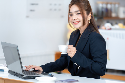 Half-body portrait. Asian female executive wearing a suit. Smile charmingly Sip coffee and trade stocks on your laptop, working in the company café zone in a relaxed and happy way.
