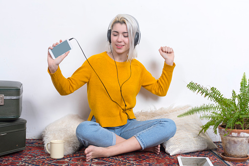 A happy woman with headphones dances while showing a smartphone, radiating enjoyment and energy in a homely, vintage-decorated space with plants.