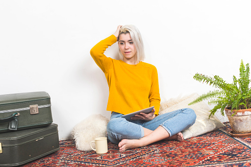 A casual woman with a tablet pauses thoughtfully, her hand in her hair, against a simple home backdrop with a vintage suitcase and lush greenery, reflecting ease and reflection.