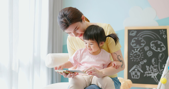 Asian mother accompany her child read story book - girl sitting on mom leg wtih safety and enjoyable