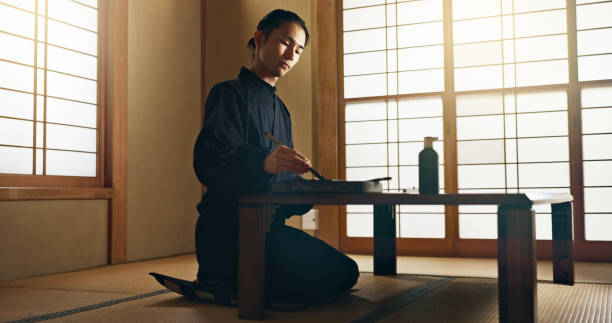 istock Man, calligraphy or creative Japanese artist in studio for art and script, letter with table, paper or alphabet. Asian, brush or person with tools, paintbrush and focus with traditional stationery 2054972728