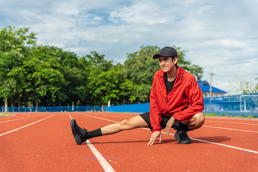 Medium shot, Young Asian man stretching his leg warm up before workout outdoor running track background. The man in sportswear exercises outside in the morning for health and wellbeing.