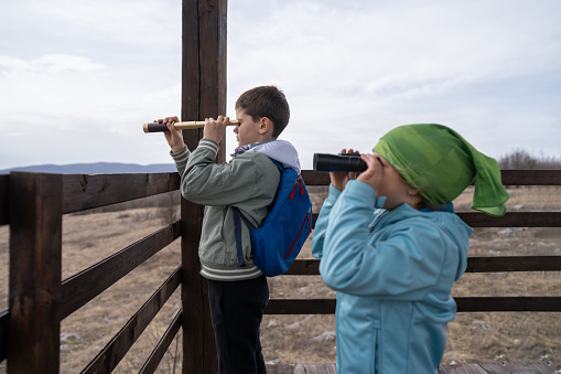 Side view of brother and little sister observing nature through telescope and binoculars from hunters seat