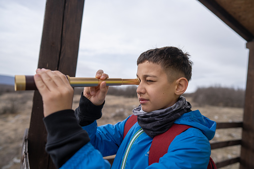 Close-up shot of boy standing on wooden hut and observing the nature through telescope on overcast day