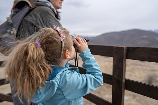 Close-up shot of little girl looking at view through binoculars while standing with her mother on the wooden hut on mountain