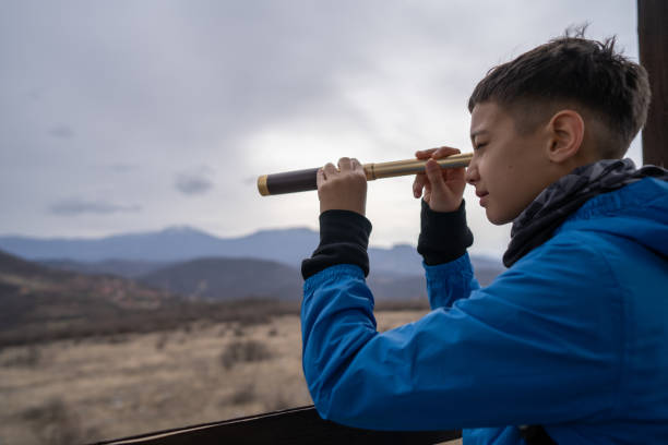 Close-up shot of boy standing on wooden hut and observing the nature through telescope on overcast day