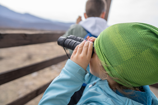 Close-up shot of little girl observing nature through binoculars with her brother from wooden hut