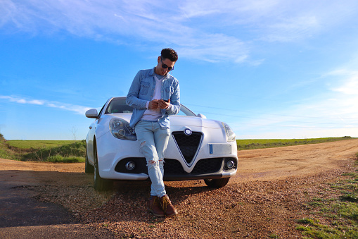 Curly haired Caucasian man leaning on his car looking at the mobile phone on a road beside the highway and the blue sky in the background, making a stop to get some fresh air. Lifestyle concept