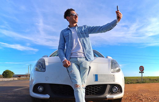 Curly haired Caucasian man leaning on his car smiling taking a selfie on the side of the road with blue sky in background. Lifestyle concept