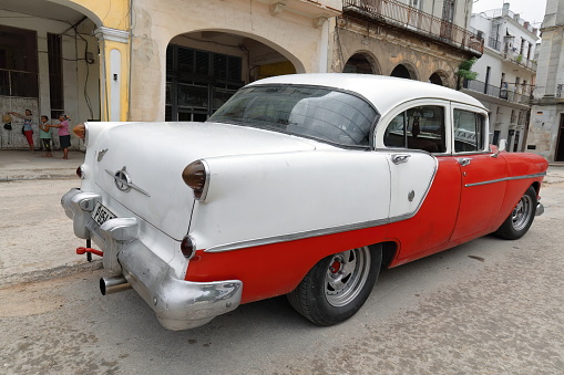 Havana, Cuba-October 07, 2019: Rd-white old American classic car -almendron, yank tank- Oldsmobile Super 88 Sedan 4 door from 1954 stops on the Plaza del Cristo Square, incidental people at doing gym.