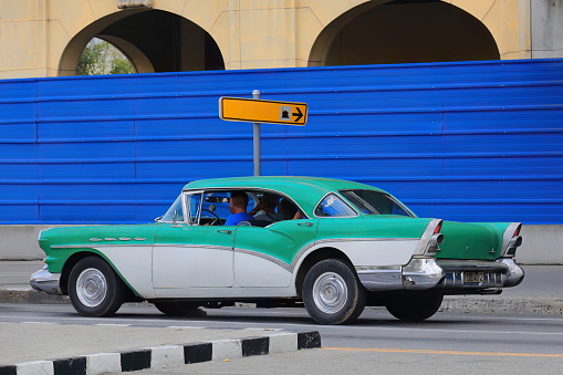 Havana, Cuba-October 07, 2019: Green-white old American classic car -almendron, yank tank- Buick Century Riviera 4 door Sedan from 1957 stops at a red light, Paseo del Prado past a sheet metal fence.