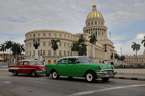 Havana, Cuba-October 07, 2019: Red-white, green-white American classic 4 door cars -almendron, yank tank- Buick Special '56, Pontiac Chieftain '57 wait at a red light on Dragones st., Capitol in back.