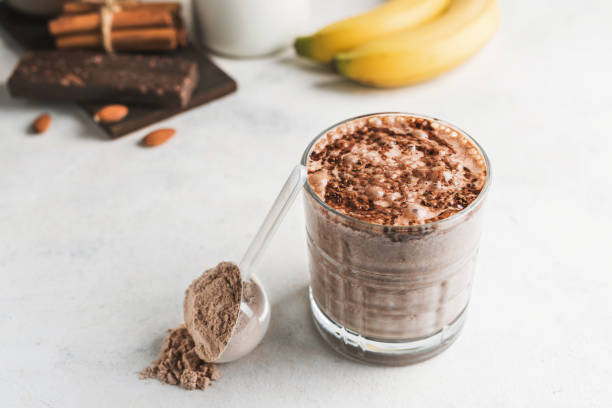 istock Glass with chocolate protein drink, milkshake smoothie on white table with bananas, protein powder in measuring spoon, protein bar, almond nuts and cinnamon sticks 2055281937