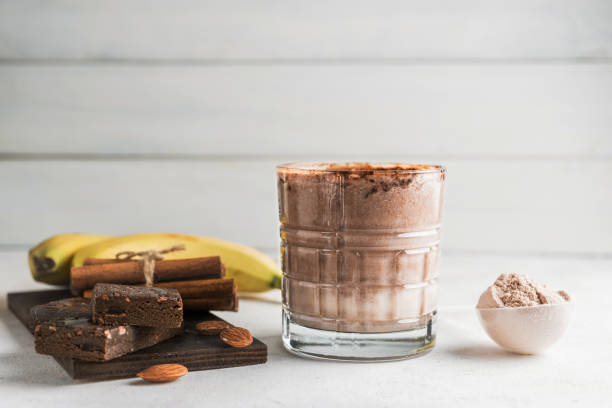 istock Glass with chocolate protein drink, milkshake smoothie on white table with bananas, protein powder in measuring spoon, protein bar, almond nuts and cinnamon sticks 2055295179