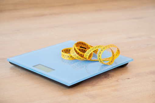 Closeup of a home scale and measuring tape on top of the blue scale on a wooden floor.
