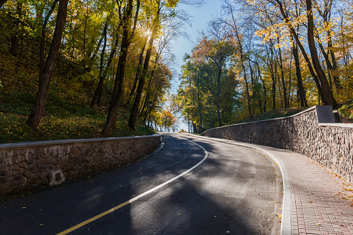 Local narrow asphalt road with sidewalk paved with stone paving tiles and stone retaining walls at the beginning of the descent in autumn forest in sunny day backlit