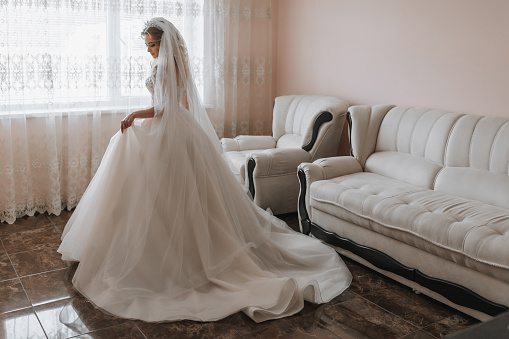 the bride in a white wedding dress. happy beautiful young woman in white traditional wedding dress in her room. large windows and light walls.