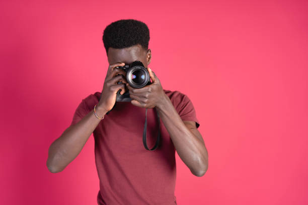 istock Young black man taking a photograph with a mirrorless camera, wearing a red t-shirt on a carmine red background with copy space. 2055409607