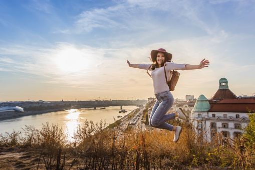 Young student woman jumping opposite famous facade and entrance to Hotel Gellert on banks of Danube in Budapest, Hungary in sunrise