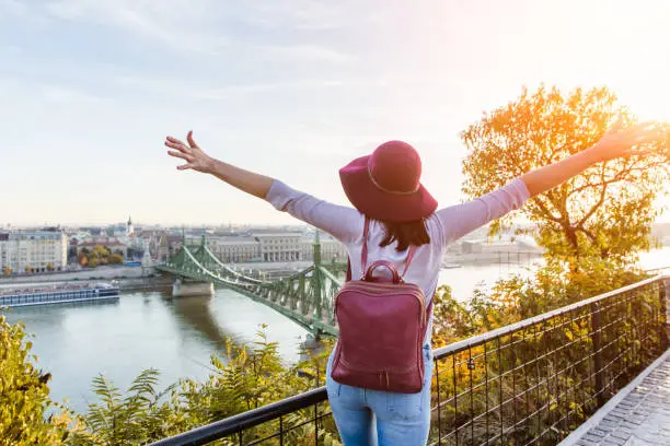 A happy young woman in purple hat enjoying her trip to Budapest, Hungary from the point from Gellert Hill during sunrise in autumn