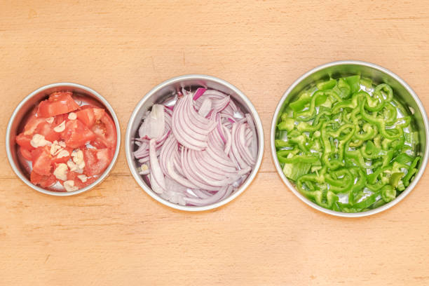 Three different sized circular metallic plates set from small to large, filled with raw tomatoes and garlics, slices onions and pieces of sliced green pepper. Separated and set on a wooden table top.