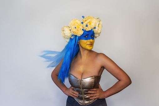 Beautiful creative young woman smiling while looking at camera on isolated background. Girl dressed in bright corset, blue hair, yellow blue face art, wind blows hair, wreath of spring flowers on head