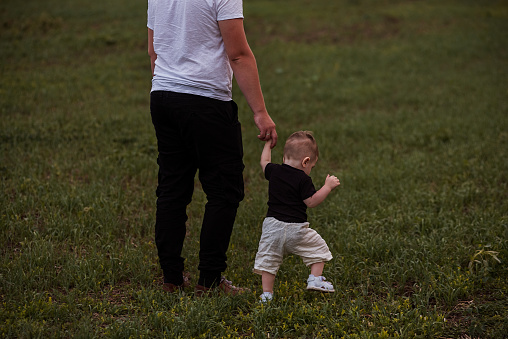 Young man holds the hand of little toddler. Father and son are walking along green field. Parental care, protection. Fathers Day. Diversity family Bonding at Dusk