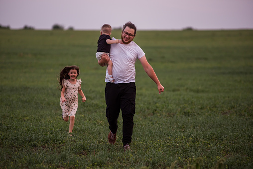 Young father runs with little son in arms and older daughter across green field. Diversity Playful Family Time in Meadow. Fathers day.