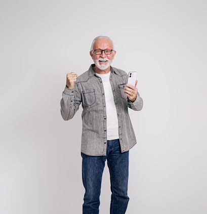 Excited old businessman with mobile phone shaking fist after reading good news on white background