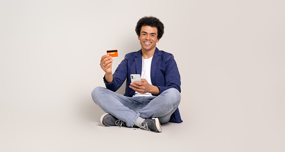 Full length of smiling businessman with mobile phone and credit card sitting over white background