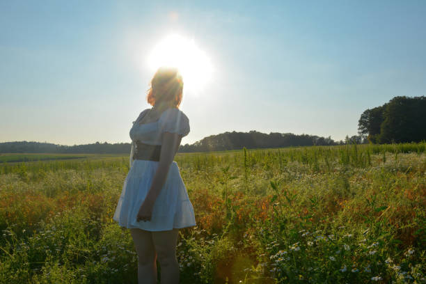 A pretty young woman from the front, wearing a white short dress, stands in green nature with the bright sun