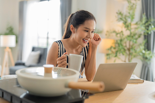 morning goodvibe lifestyle of moden simple living  asian female woman enjoy coffee and update news or social trend with laptop media newspaper while hand hold coffee stay in kitchen interior home background