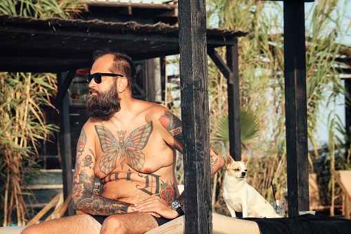 Tattoed bearded hipster looking guy relaxing on a beach with his apple head chihuahua