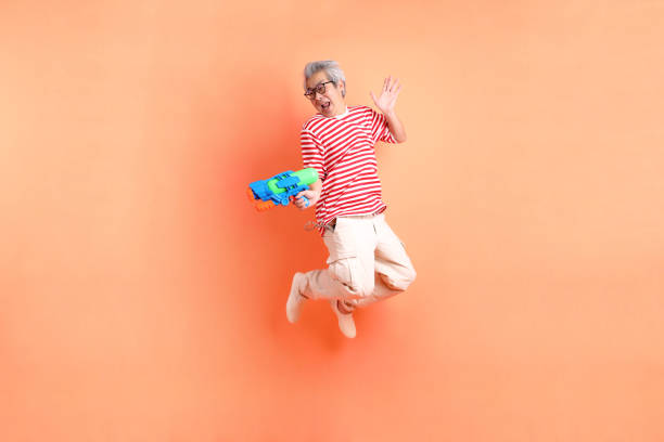 HAPPY SONGKRAN DAY. Asian energetic senior man in STRIPED TOPS and eyesglasses with gesture of holding plastic water gun isolated on orange background. Songkran festival. Thai New Year's Day.