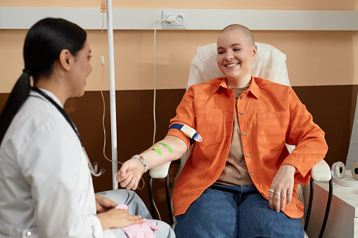 Portrait of smiling bald woman receiving chemotherapy treatment in procedure room at clinic and talking to nurse