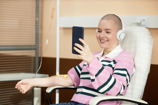 Portrait of smiling bald woman receiving IV treatment in clinic and talking via video chat copy space