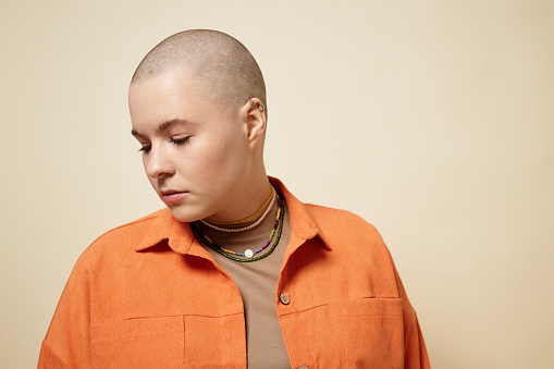 Minimal portrait of bald young woman wearing bright orange jacket looking away in studio, copy space